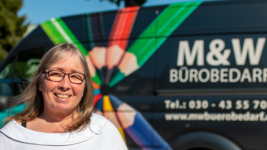 Birgit steht mit strahlendem Lächeln vor einem ihrer Transporter. Dieser trägt die Aufschrift M&W Bürobedarf und ist mit Buntstiften verziert. Birgit hat blondes Haar trägt eine Brille und eine weiße Bluse.