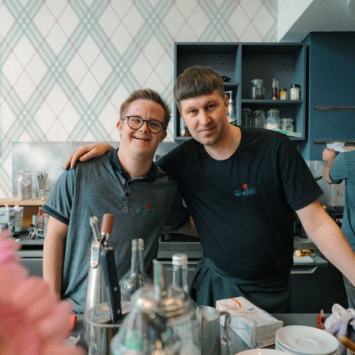 Zwei Mitarbeitende, einer mit erkennbarer Behinderung, stehen hinterm Tresen des Cafés und lächeln Arm in Arm in die Kamera.
